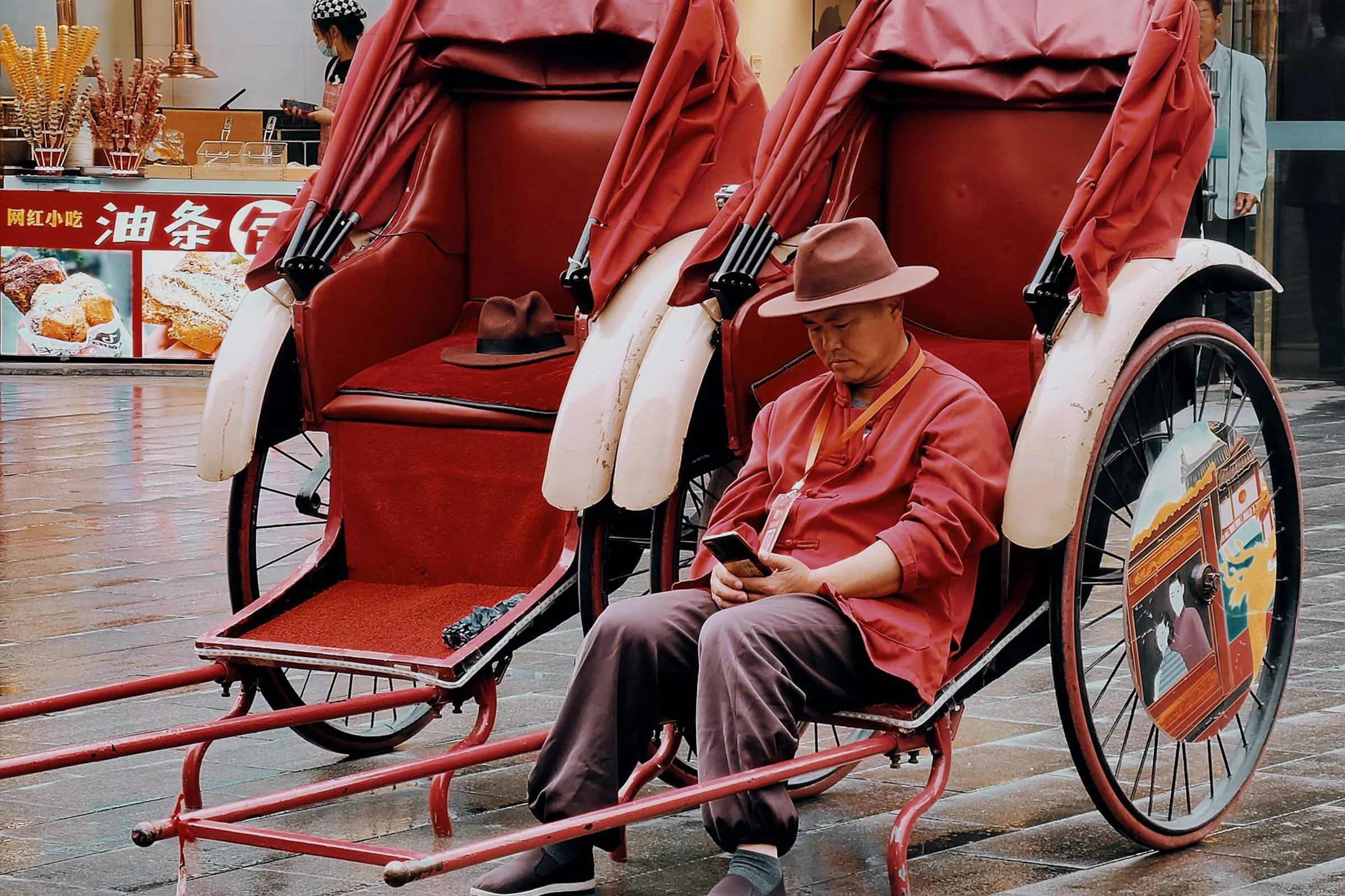 A rickshaw driver in red using a mobile phone on a Nanjing street, with Chinese food stalls in the background