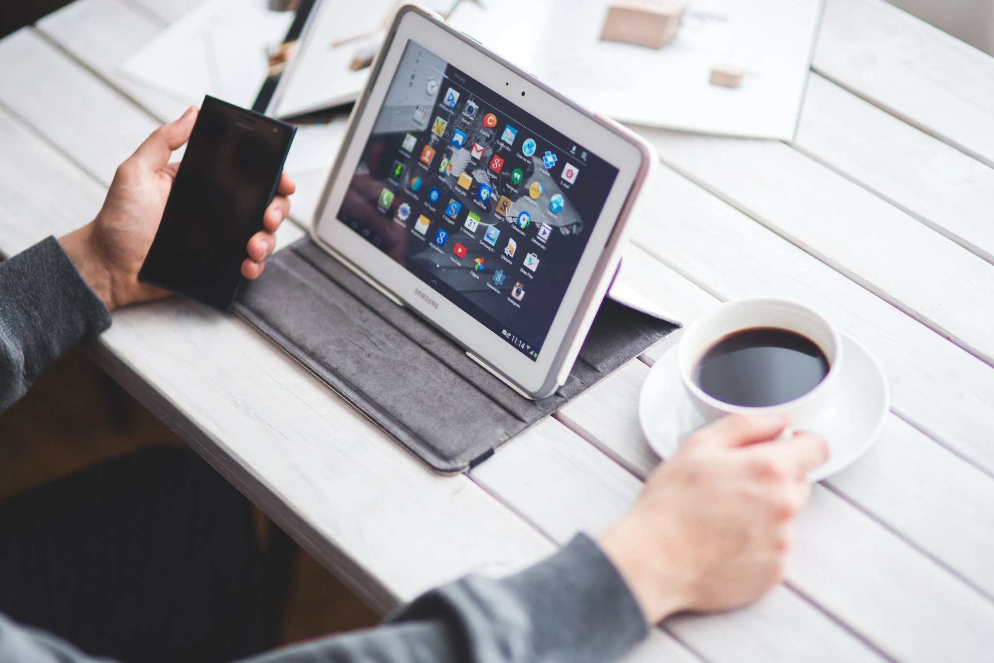 A hand holding a phone next to a tablet propped up on a white wooden cafe table beside a cup of black coffee, set up for remote work