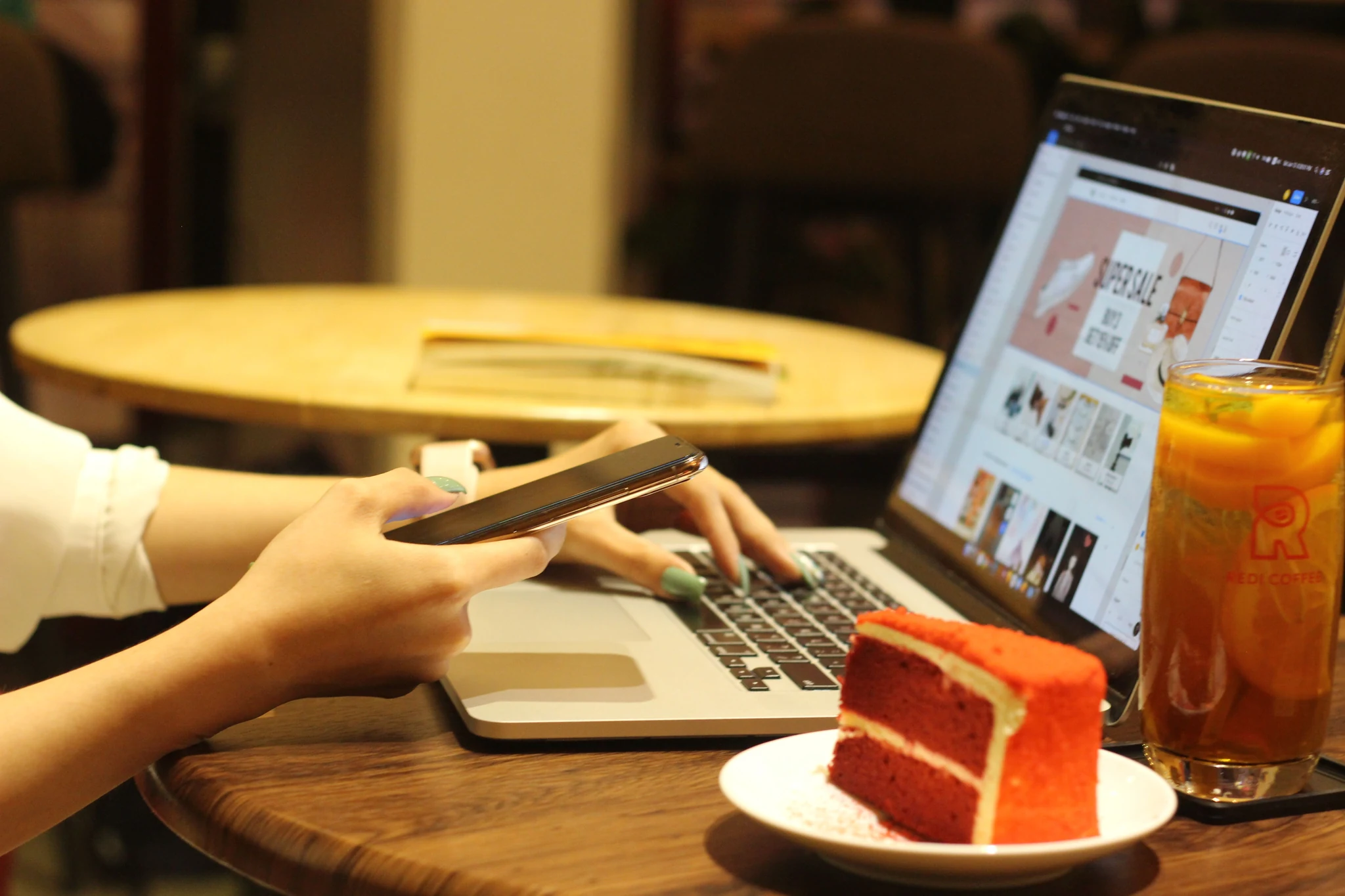 A hand scrolling a smartphone above a laptop keyboard at a cafe table, with a slice of red velvet cake and a tall glass of iced fruit tea beside it