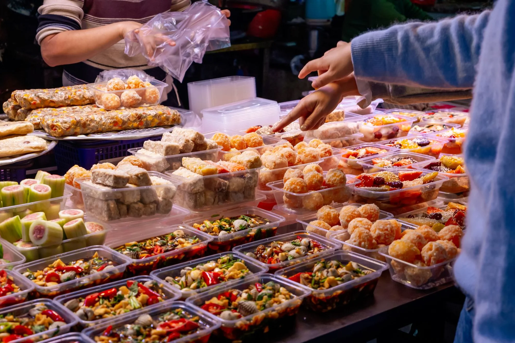 A market stall in China with rows of plastic takeout containers filled with prepared dishes, with a customer reaching for an item