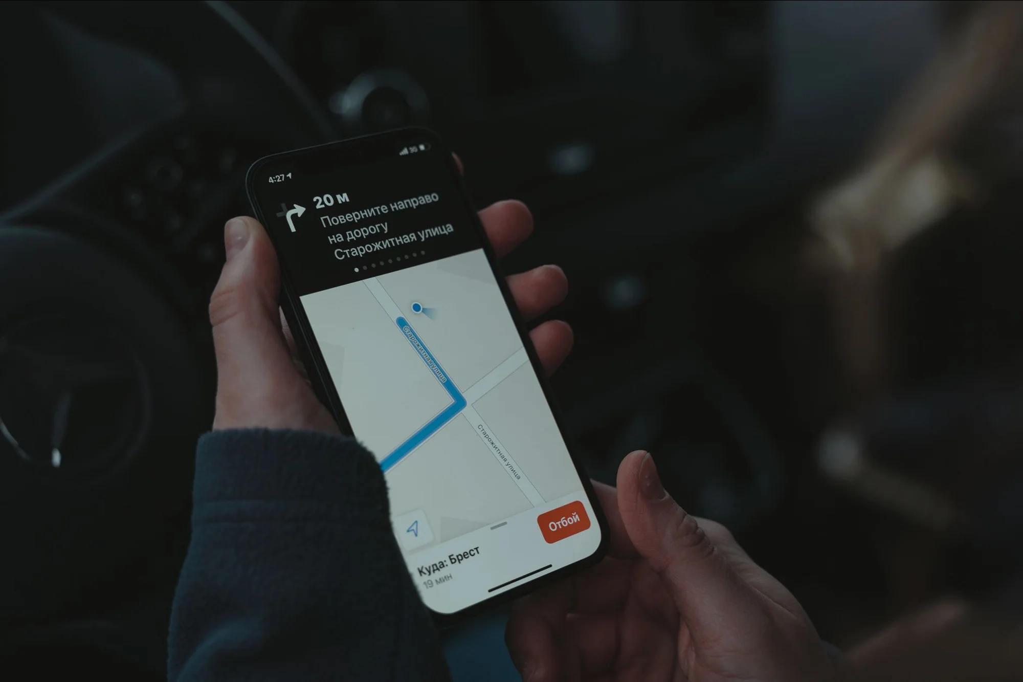 A hand holding a phone displaying a maps app turn-by-turn navigation screen in a car