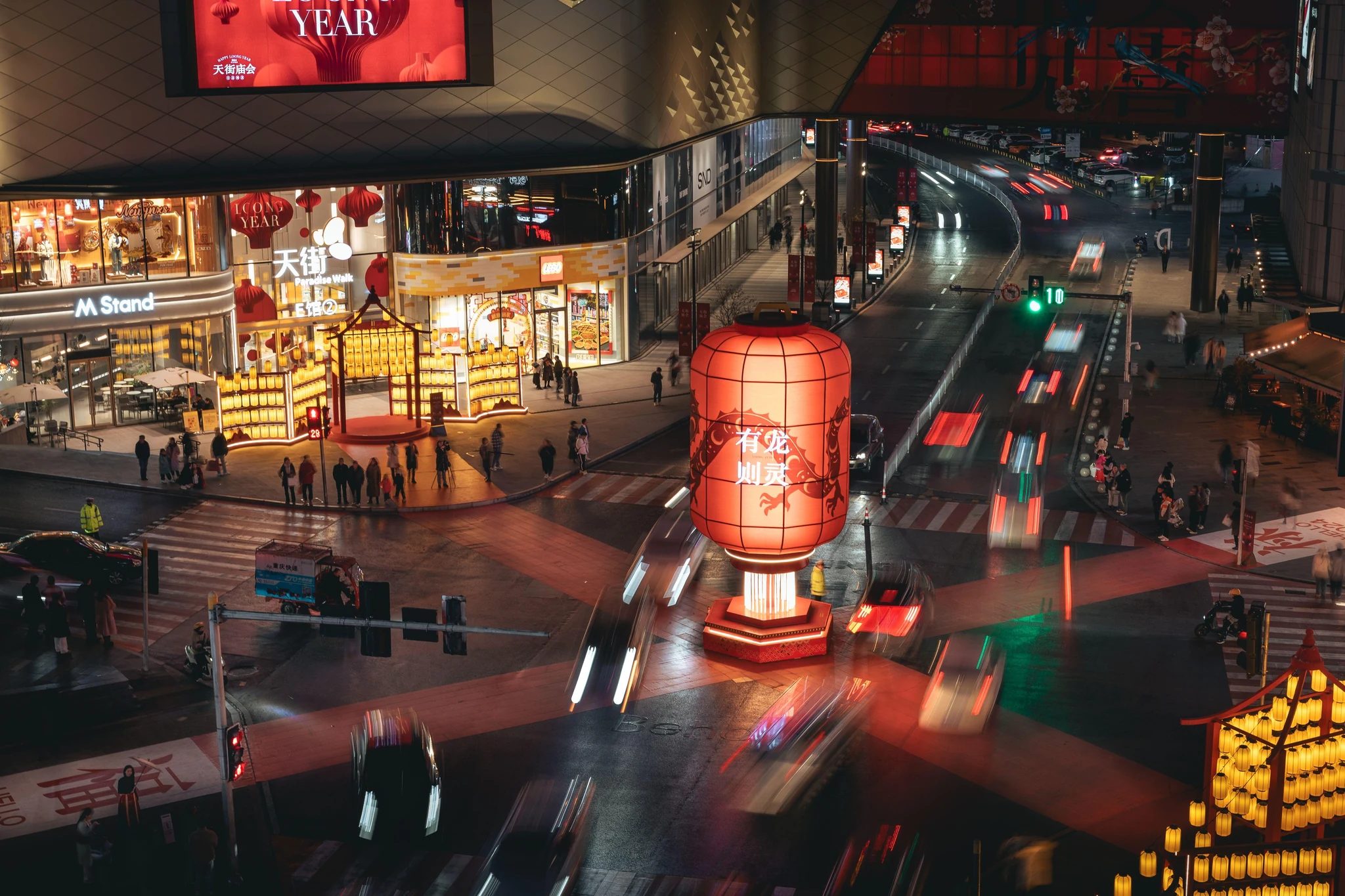 A well-lit Chinese commercial pedestrian street at night with red lanterns, storefronts and light trails from passing traffic