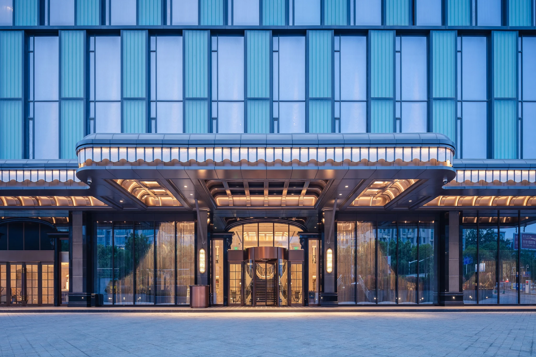 The glass-fronted entrance of a well-lit Chinese hotel with a porte-cochere and revolving door at dusk