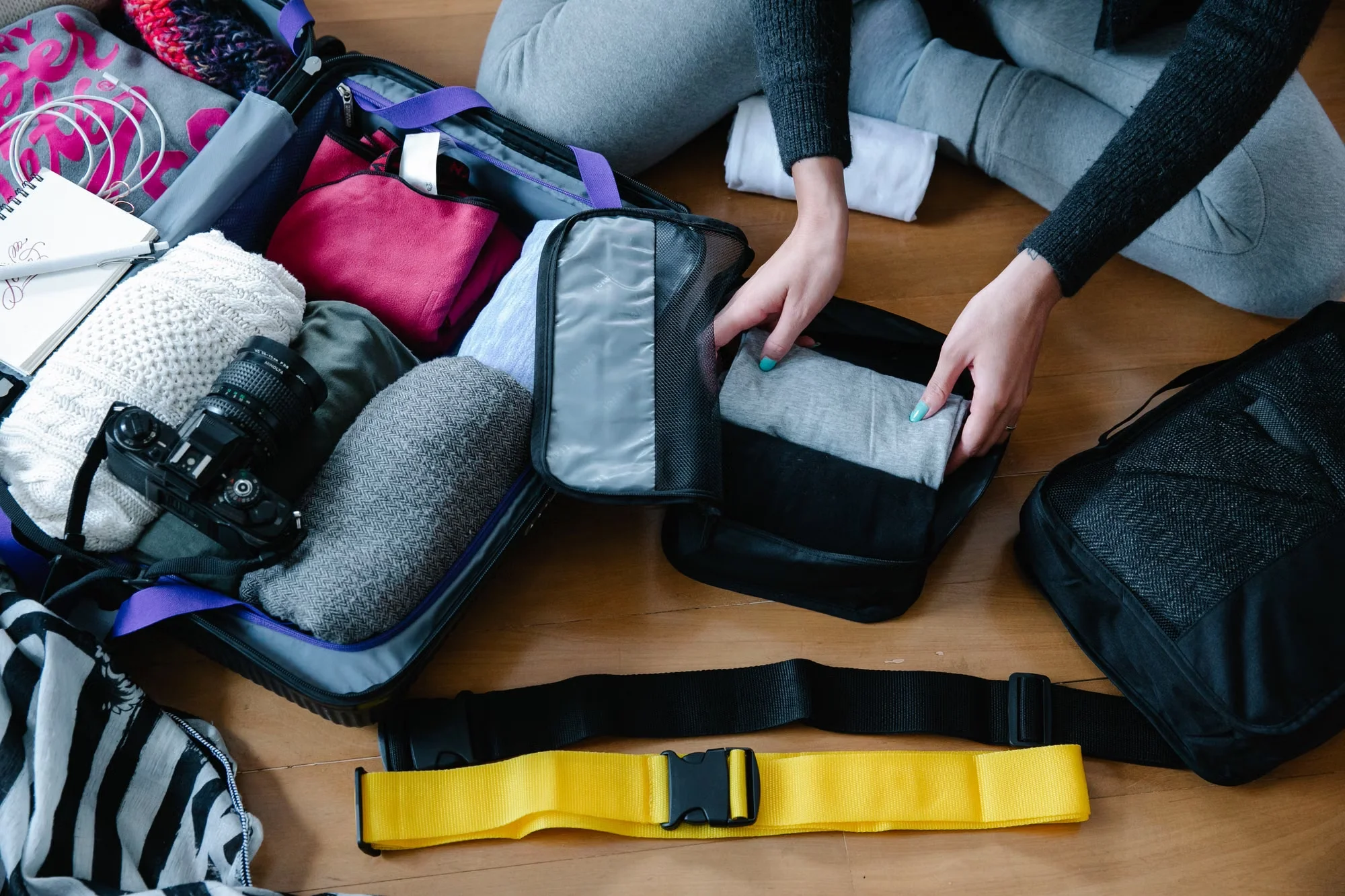 A traveler packing a suitcase on a wooden floor with clothes, a camera and packing cubes spread around