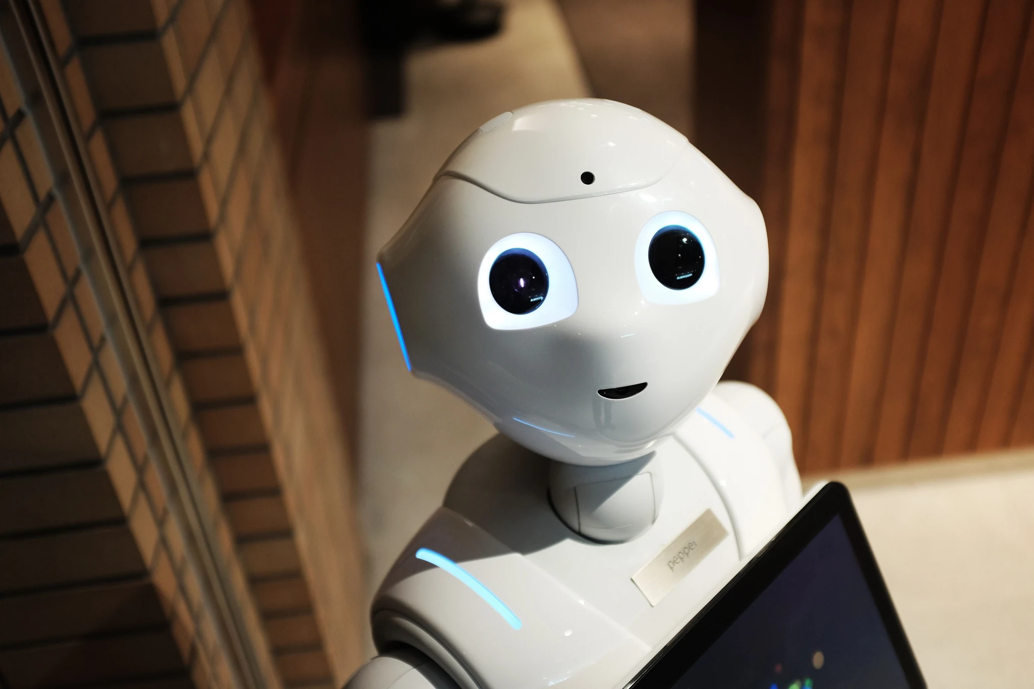 A white humanoid Pepper robot with two glowing blue eyes standing in a hotel lobby next to a tablet screen, looking straight ahead