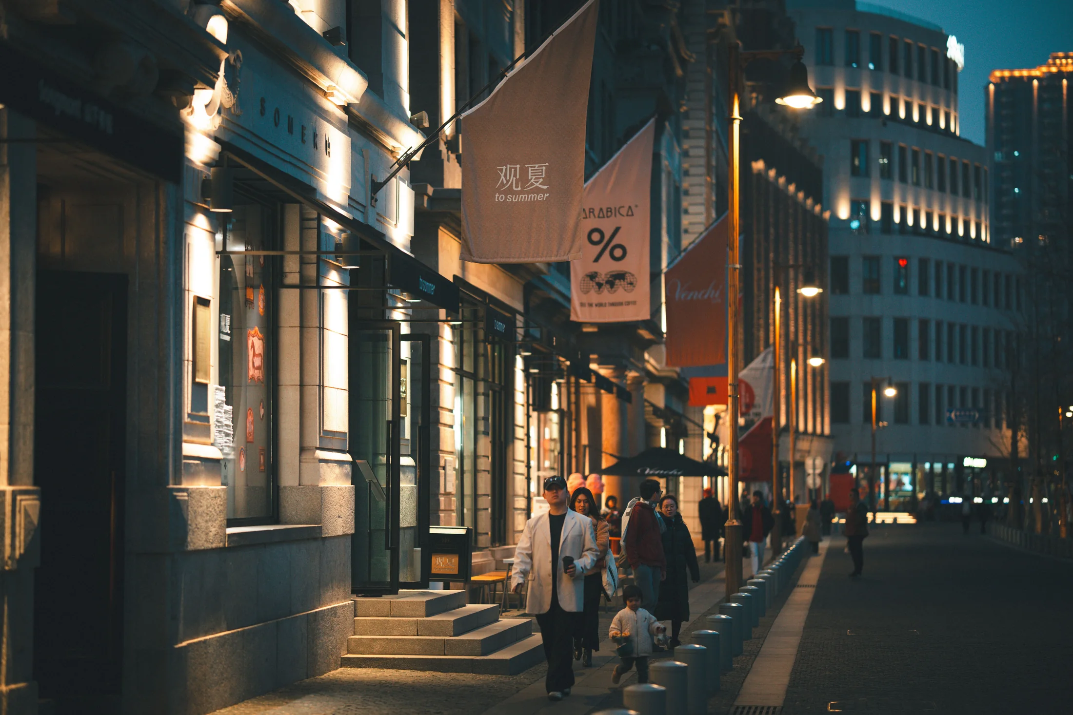 A wide evening street on the Shanghai Bund: art-deco facades lit warm, a large warm orange To Summer 'to summer' lantern and an % Arabica storefront cloth banner hanging above the sidewalk, with a few pedestrians walking under the building lights.