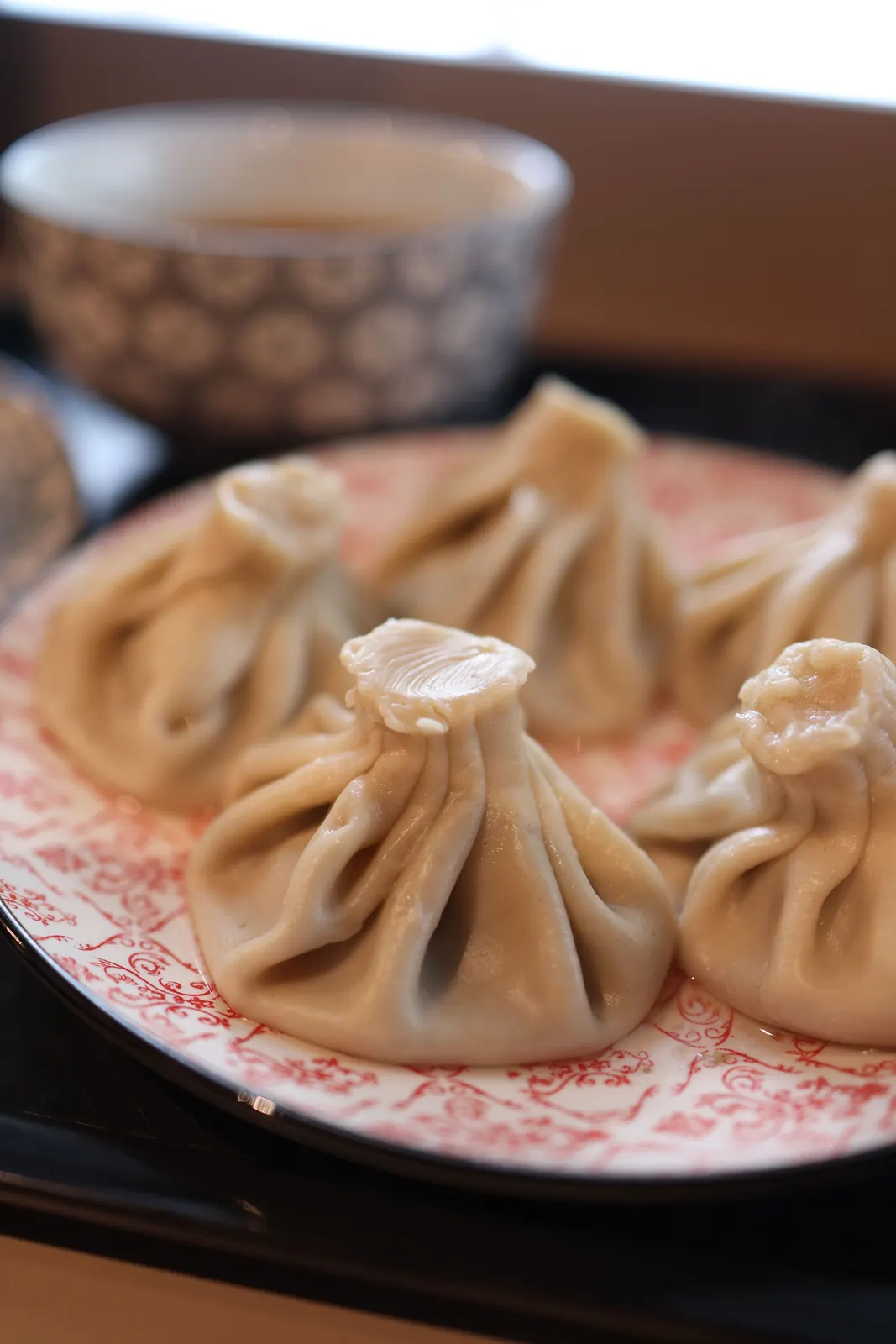 A plate of steamed xiaolongbao soup dumplings with twisted tops, served on a traditional patterned plate with a dipping bowl in the background.